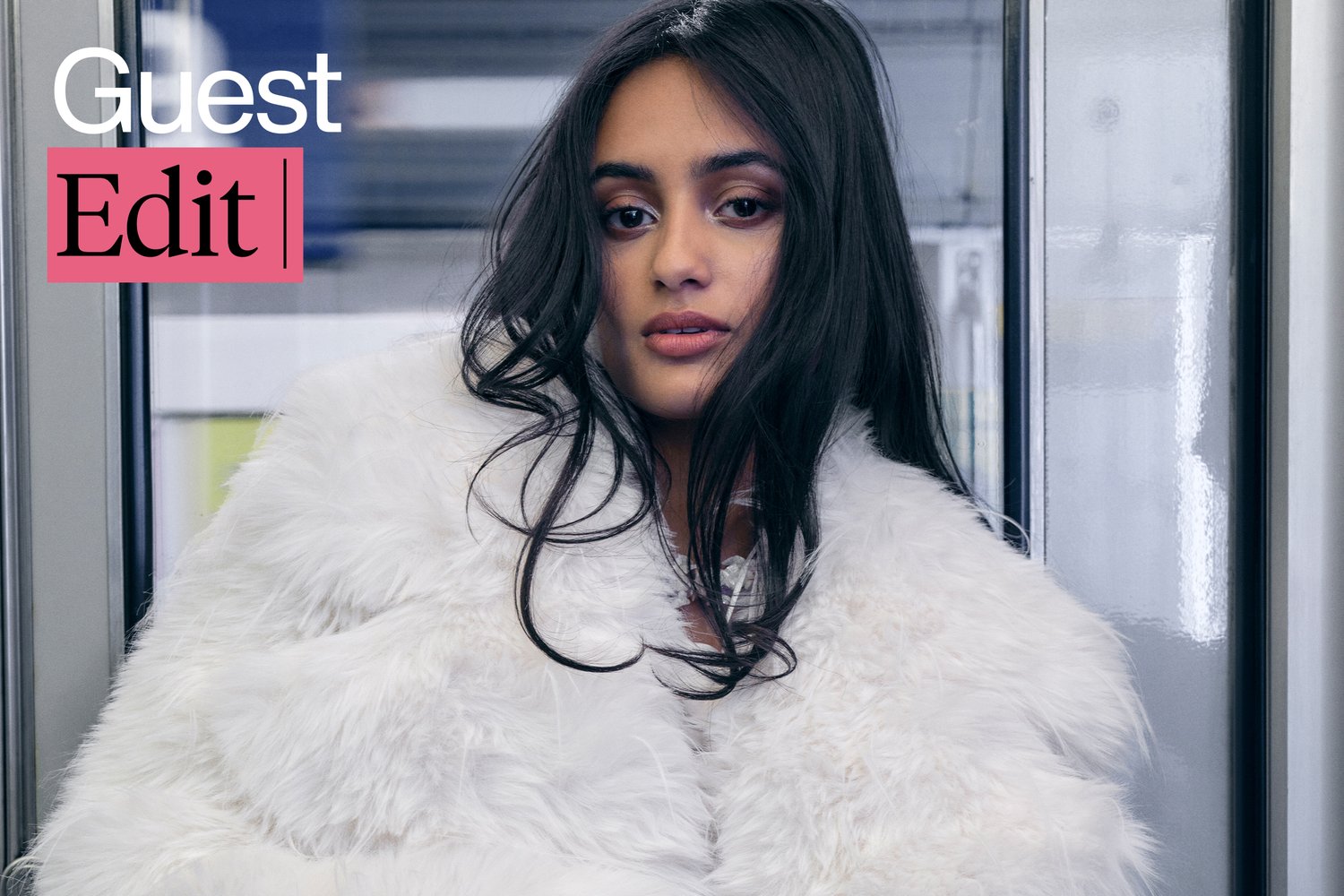 A woman with long dark hair, wearing a fluffy white coat, gazes thoughtfully at the camera in a modern indoor setting.