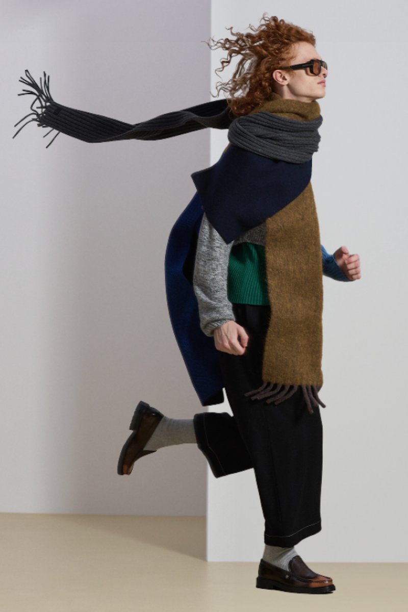 A model with curly red hair runs, wearing layered outfits including a green sweater, oversized scarf, and sunglasses, set against a minimal background.