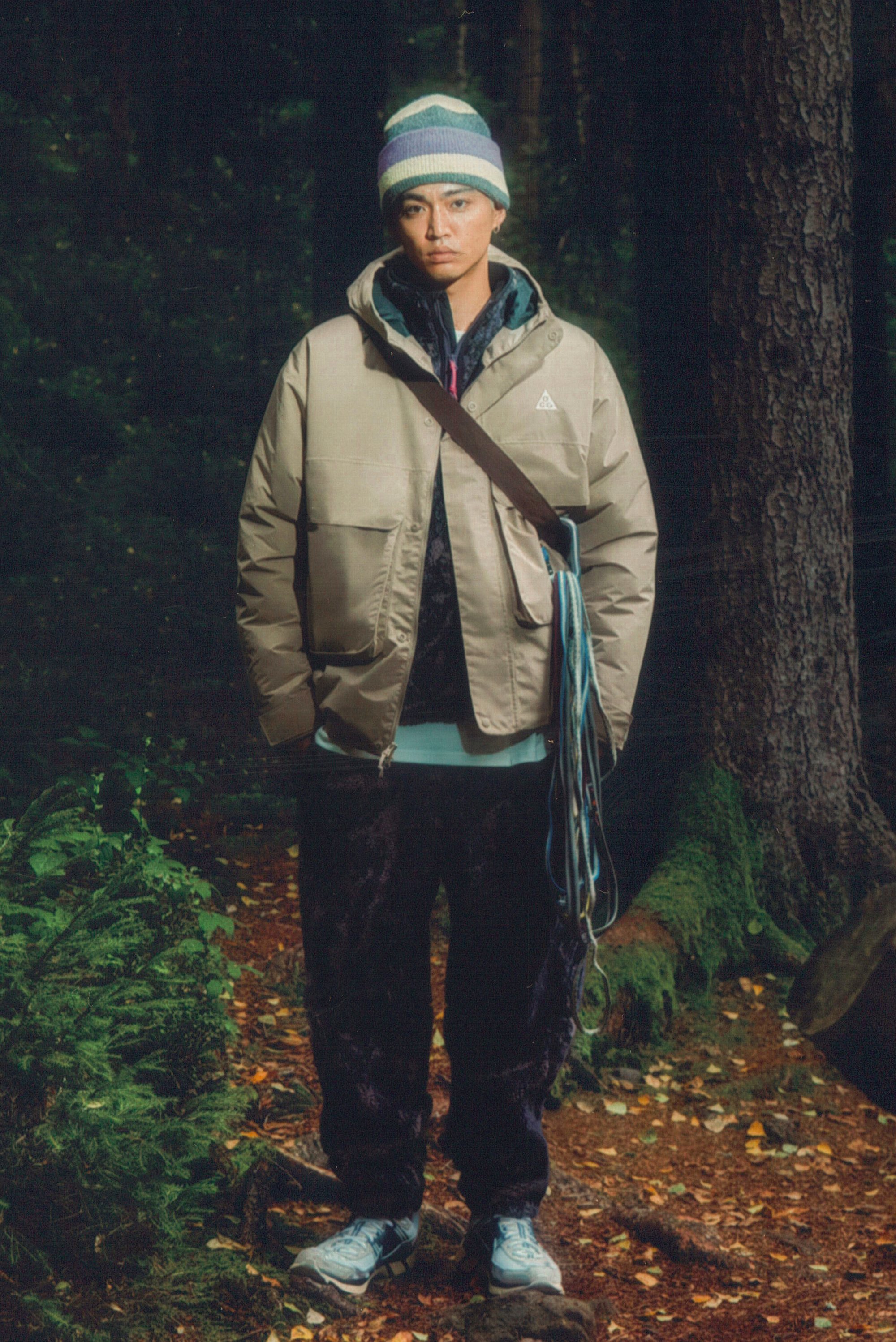 A person stands in a forest, wearing a beige jacket, dark pants, and a striped beanie, with a bag and ropes across their body.