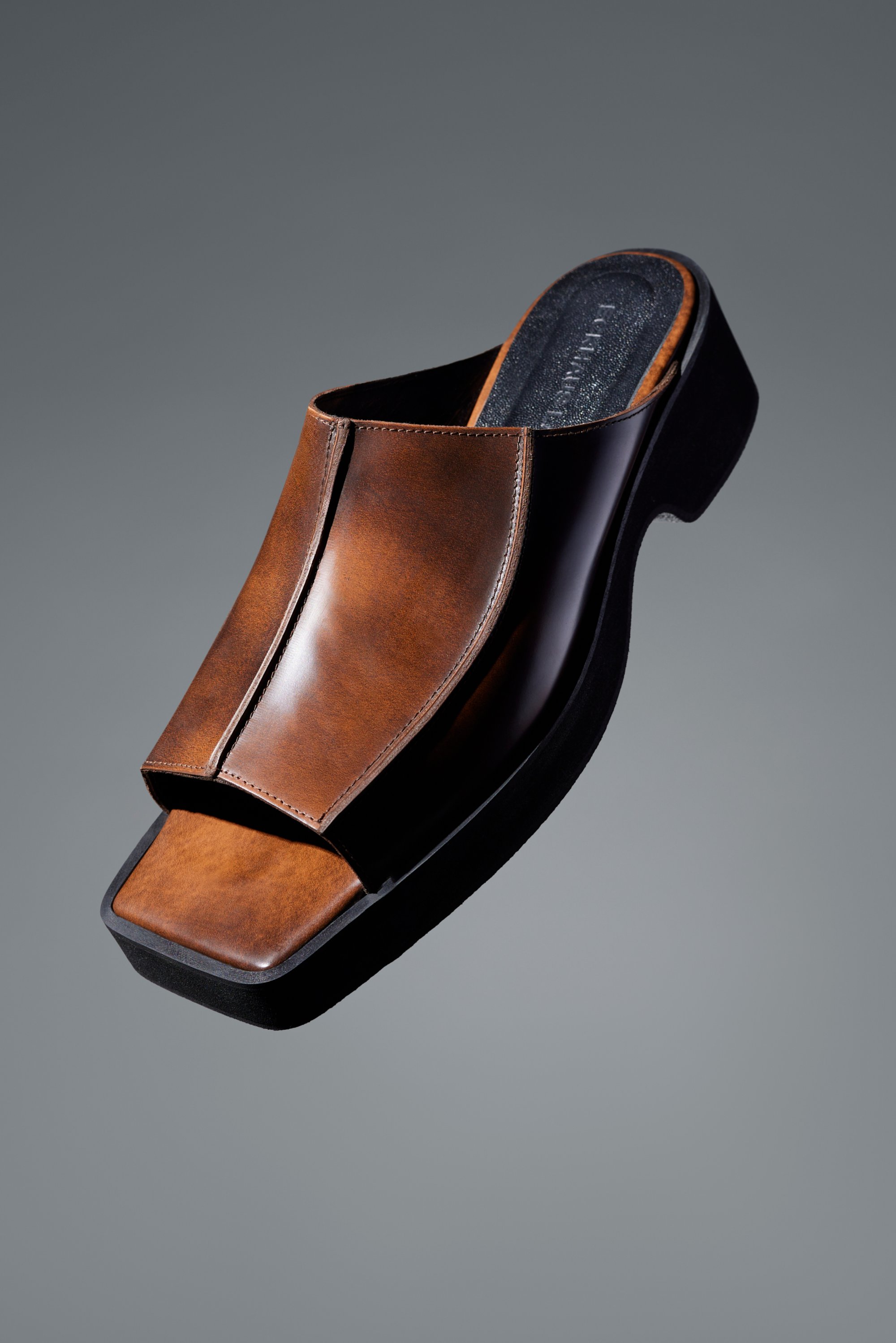 Brown leather open-toe sandal with a sleek design and a flat, black sole, positioned against a gray background.