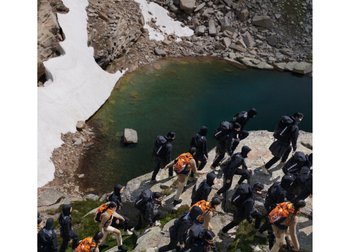 A group of hikers in black and orange gear ascends rocky terrain near a greenish lake, with patches of snow visible in the background.