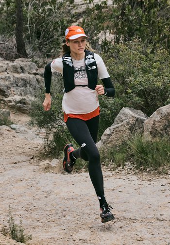 Woman wearing an orange cap and black leggings trail running on a rocky path surrounded by bushes and rocks.