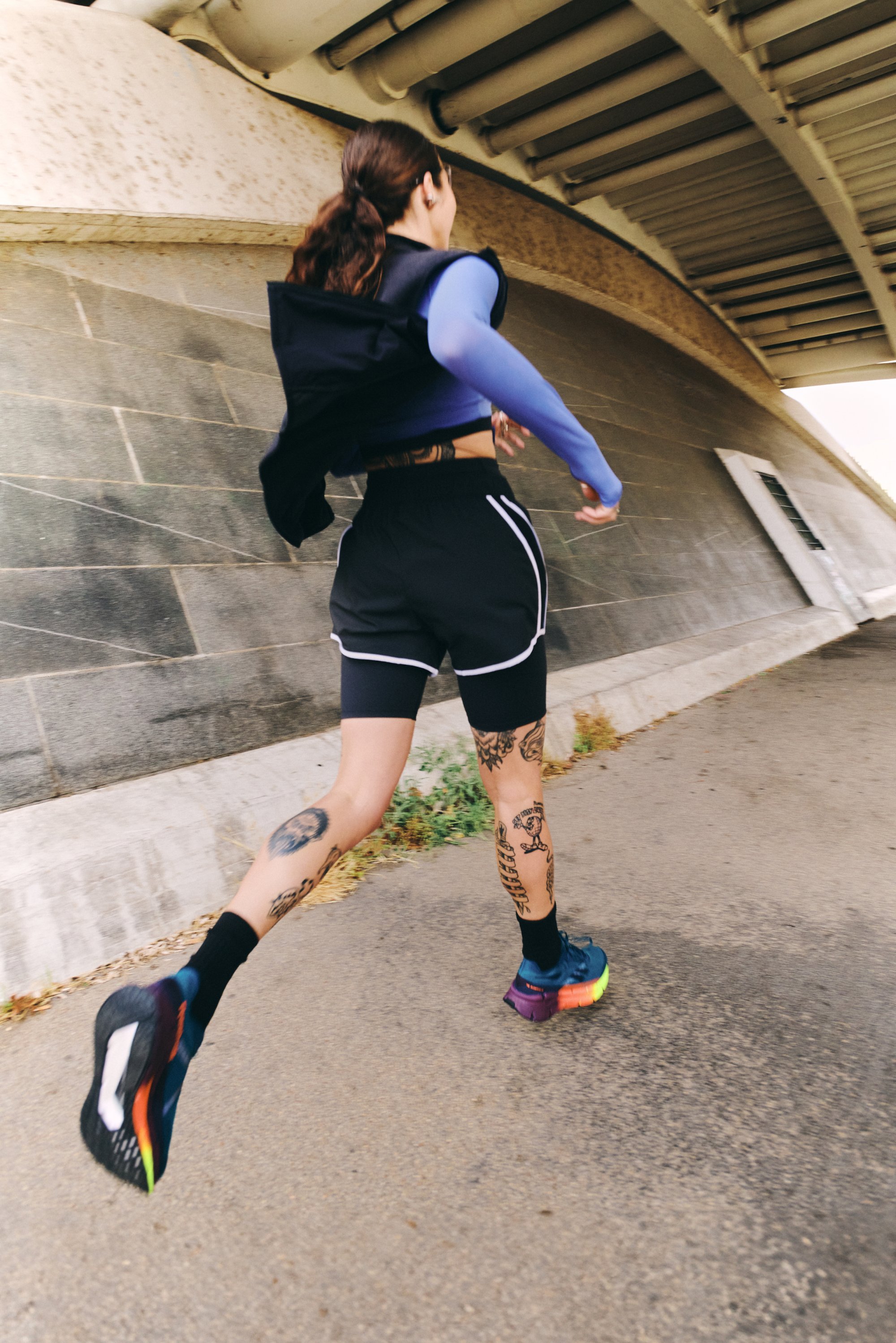 A person with tattoos runs along a pathway under an overpass, wearing a blue long-sleeve top, black shorts, and colorful sneakers.