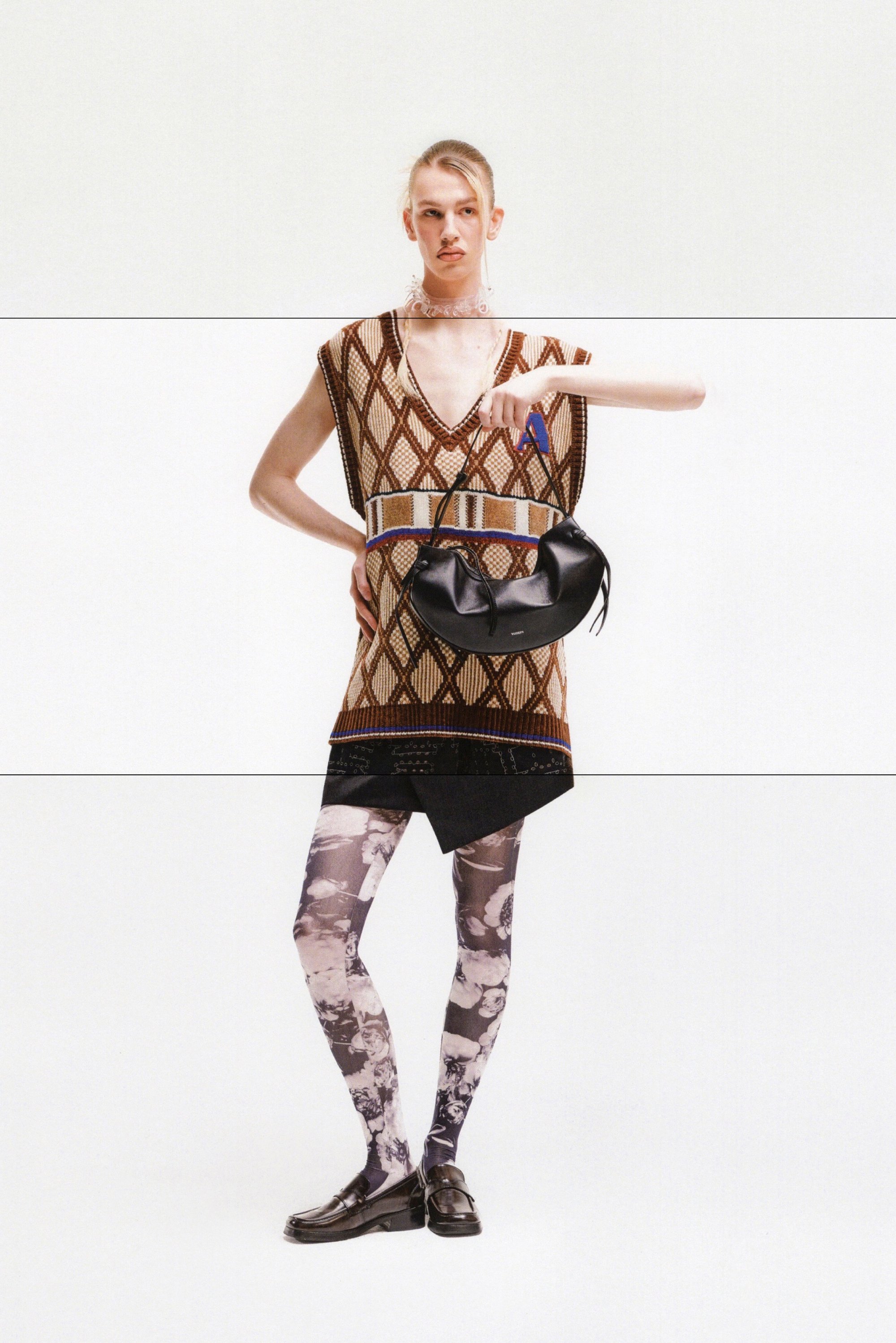 Una persona posa con una blusa sin mangas de estampado, pantalones cortos oscuros y leggings florales, sosteniendo un bolso negro y señalando con confianza.