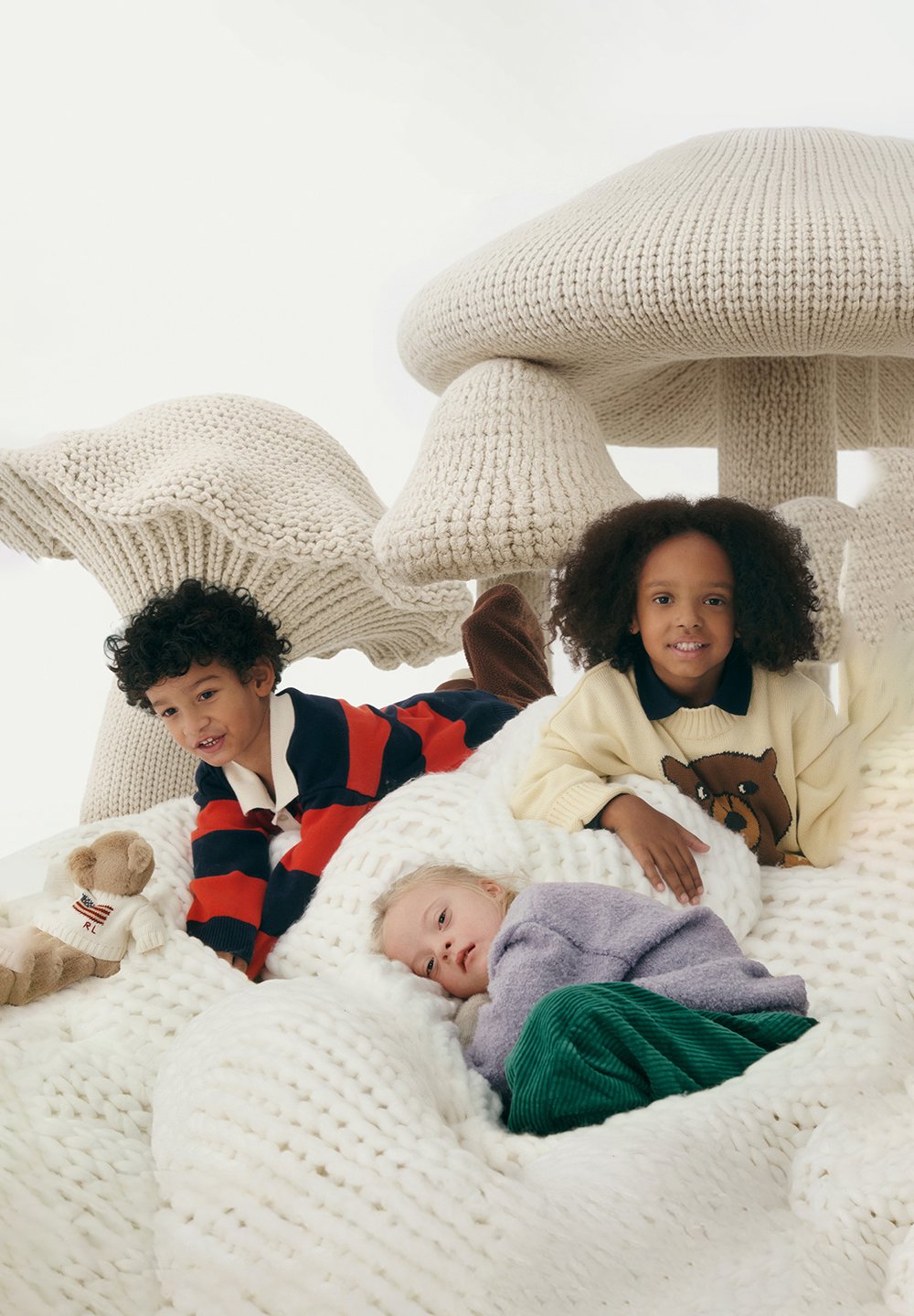 Τρία παιδιά χαλαρώνουν πάνω σε μια μεγάλη, λευκή κουβέρτα με χοντρές πλέξεις και ανάγλυφες υφές. Στο φόντο διακρίνονται υπερμεγέθεις, πλεκτά σχήματα μανιταριών σε εκρού χρώμα.