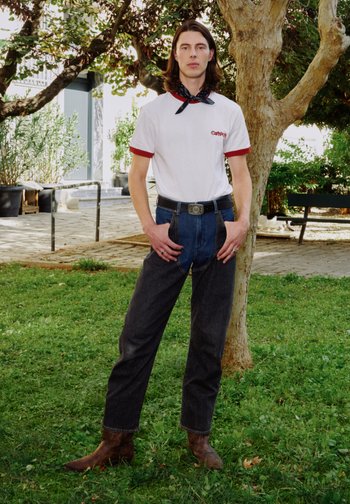 Jeune personne aux cheveux mi-longs portant un T-shirt blanc, un jean bicolore, des bottes marron et un foulard noir autour du cou, debout sur l'herbe près d'un arbre.
