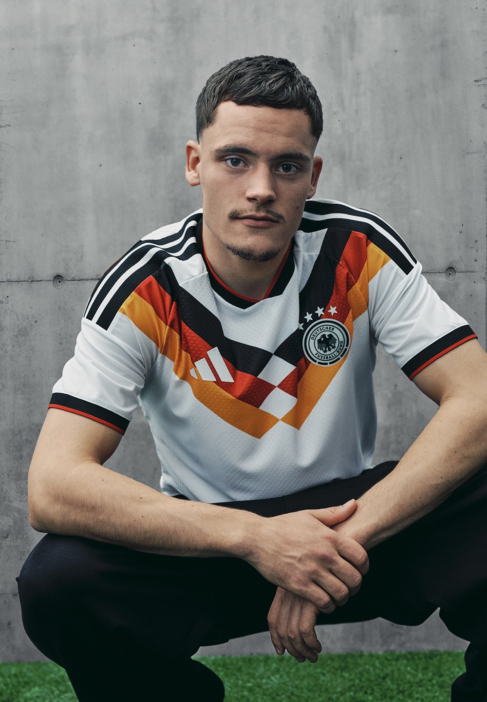 Biała koszulka piłkarska z geometrycznym wzorem w kolorze czerwonym, czarnym i żółtym, z trzema czarnymi paskami na ramionach oraz okrągłym dekoltem.