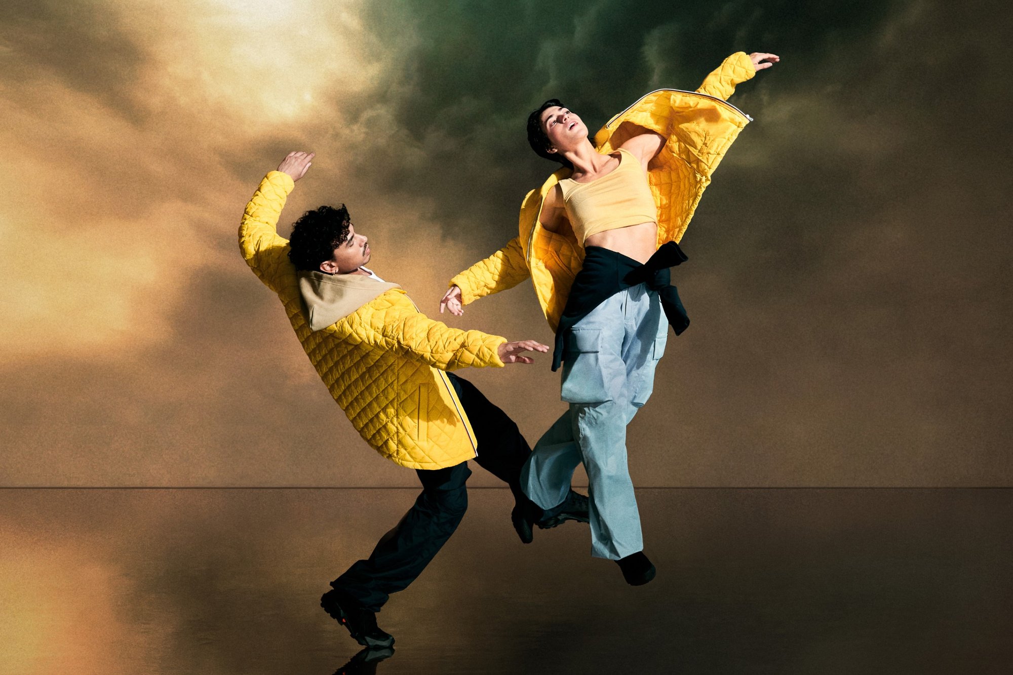 Due ballerini in giacche gialle e abbigliamento casual eseguono pose dinamiche contro uno sfondo drammatico e nuvoloso, riflettendo movimento ed energia.