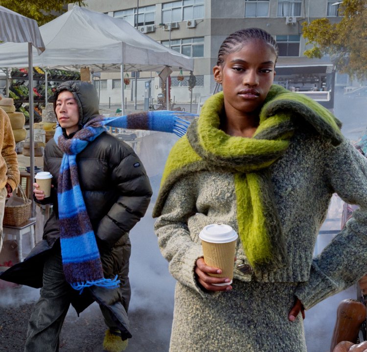 Cuatro individuos caminan por un mercado. Llevan una mezcla de abrigos grandes en tonos de marrón, negro, verde y gris, con bufandas texturizadas.