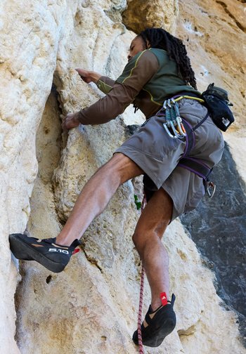 Man wearing climbing gear and shoes climbs a steep, textured rock face using handholds and footholds.