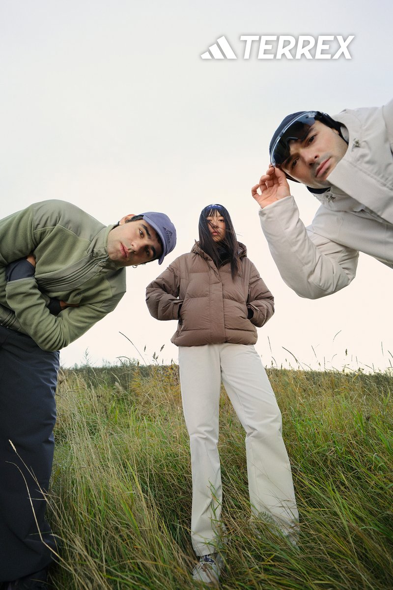 Tre personer bär utomhuskläder: en brun täckjacka, grå fleecetröja och vita byxor. Scenen visar högt gräs och en molnig himmel.