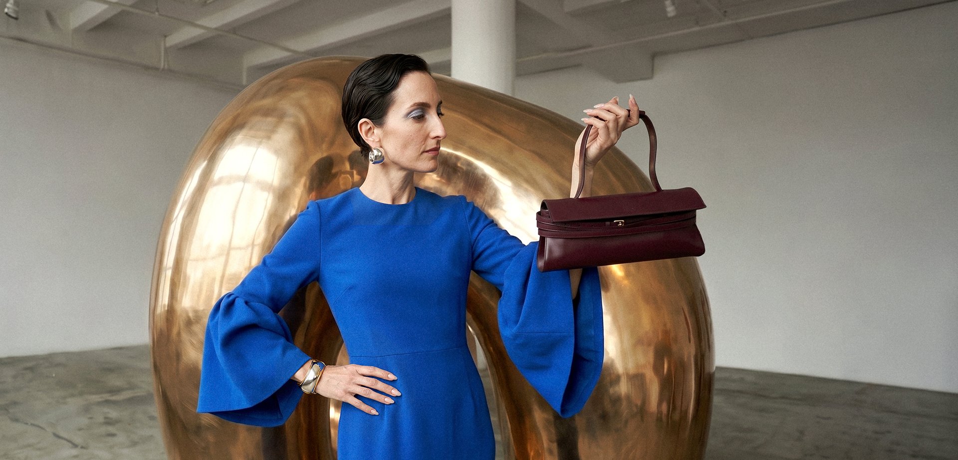 Frau in blauem Kleid hält eine dunkelrote Handtasche und steht vor einer grossen abstrakten Goldskulptur in einer geräumigen Galerie.