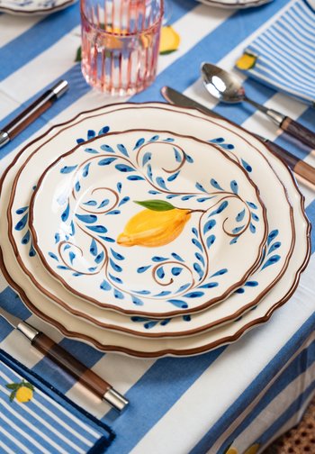 Trois assiettes en céramique décoratives avec des motifs de feuilles bleues et un design de citron jaune empilées sur une nappe rayée bleu et blanche.