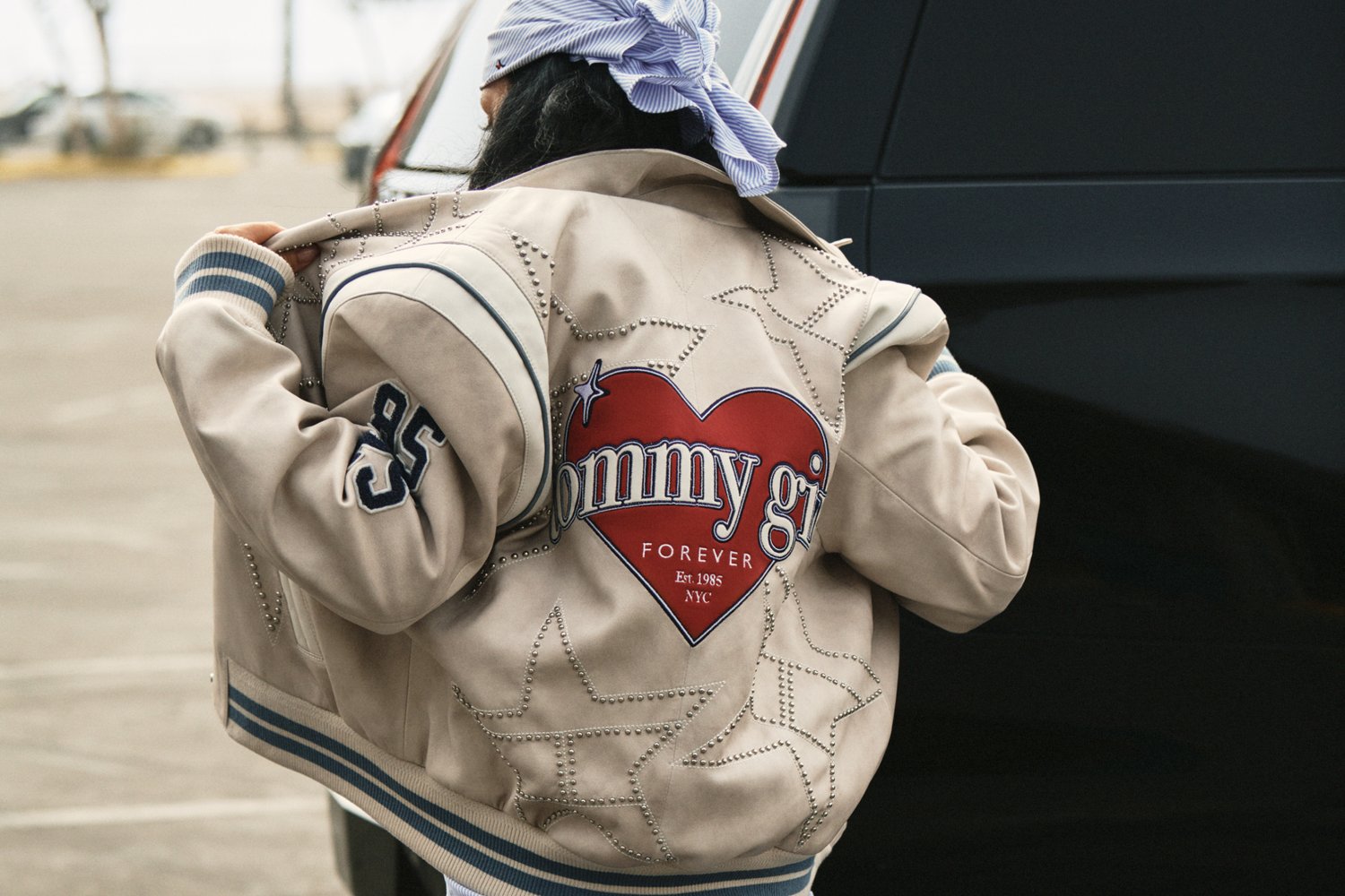 Een persoon in een beige bomberjack met "tommy girl" in een hartontwerp op de rug staat naast een zwart voertuig in een buitenomgeving.