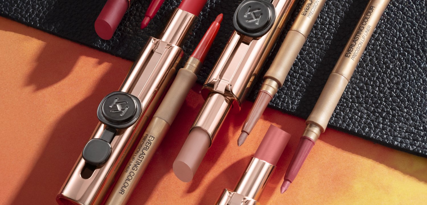 Diverse rossetti e matite labbra aperti in confezioni color oro rosa disposti su una superficie arancione e nera strutturata.