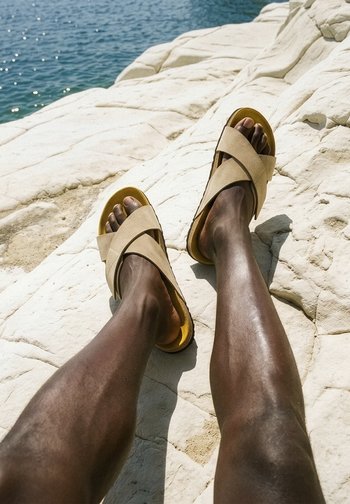Jambes à la peau foncée portant des sandales beige à brides croisées, reposant sur un rocher blanc à côté d’une eau bleue scintillante.
