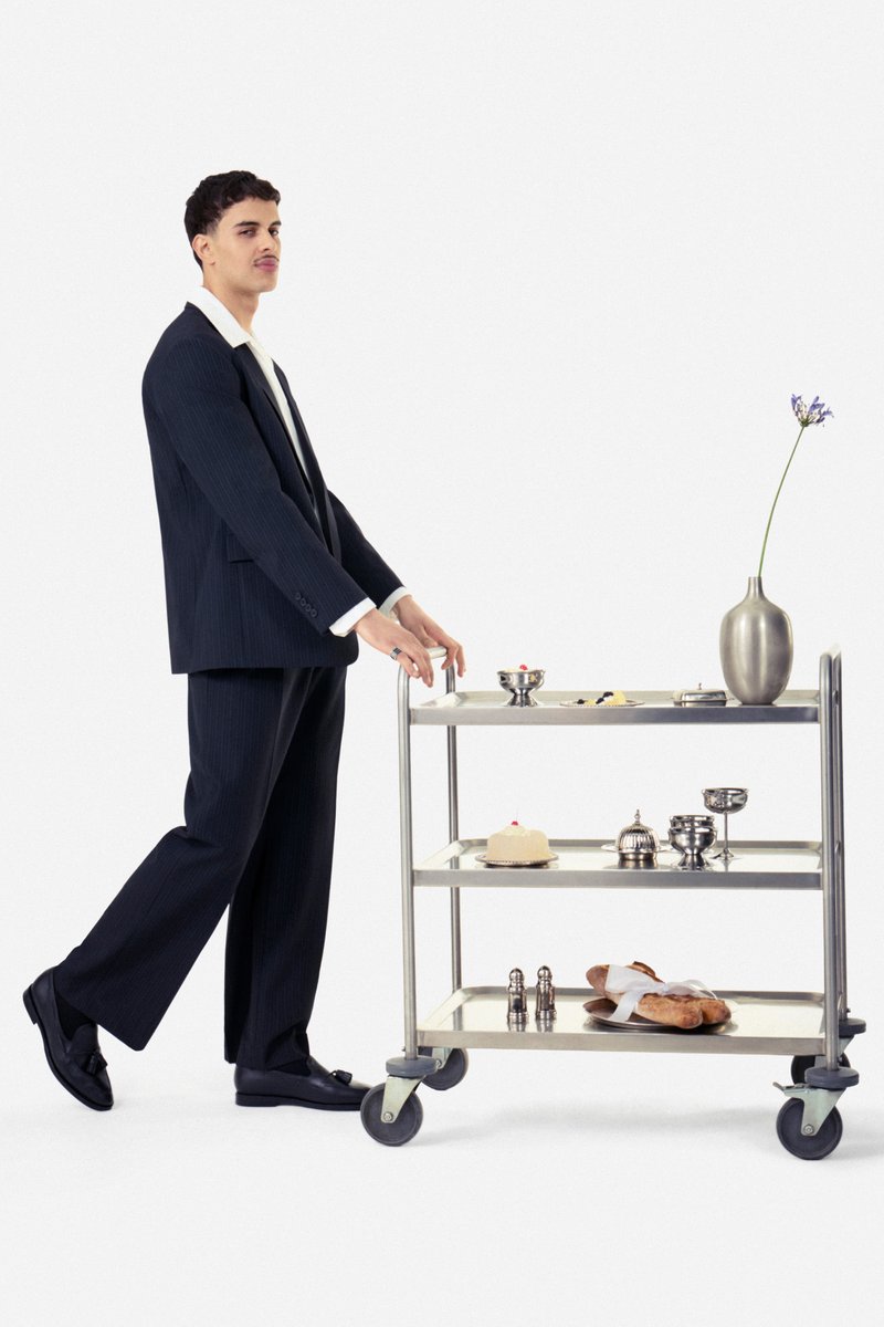 Mann in schwarzem Anzug und weißem Hemd schiebt einen Metallwagen mit Gebäck, Besteck und einer Vase mit einer einzelnen Blume vor weißem Hintergrund.