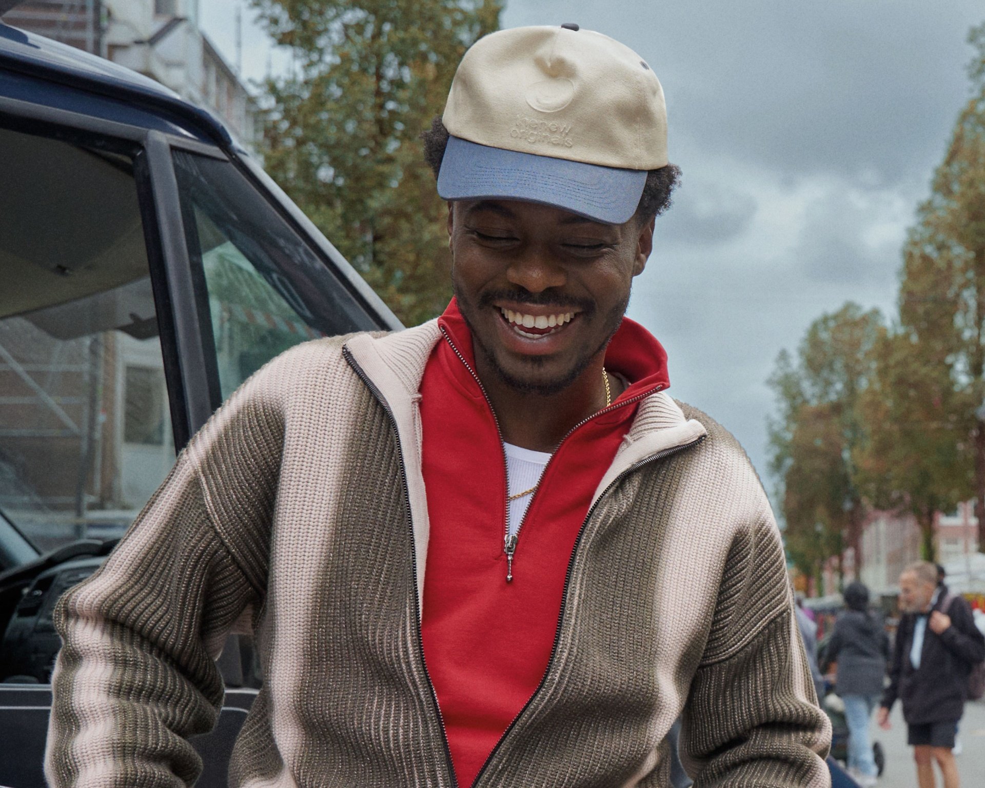 Casquette beige à visière bleue, pull en maille beige avec texture cannelée, haut rouge, silhouette souriante dans un cadre urbain en extérieur avec des arbres.