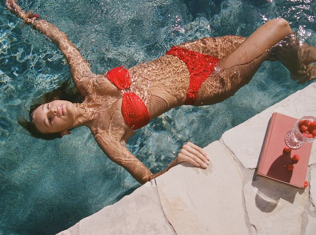 Frau in rotem Bikini, die entspannt in einem Pool treibt, hält sich am Rand fest, mit einem Buch und einem Glas mit roten Beeren am Poolrand.