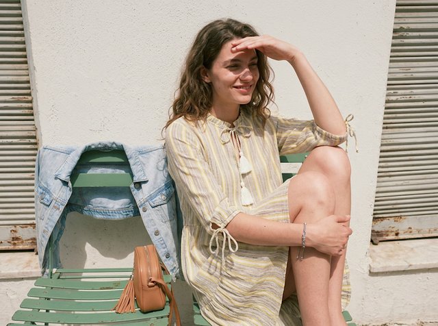 Jeune femme en robe jaune rayée assise sur un banc vert, se protégeant les yeux de la lumière du soleil, avec une veste en jean et un sac beige à côté d'elle.