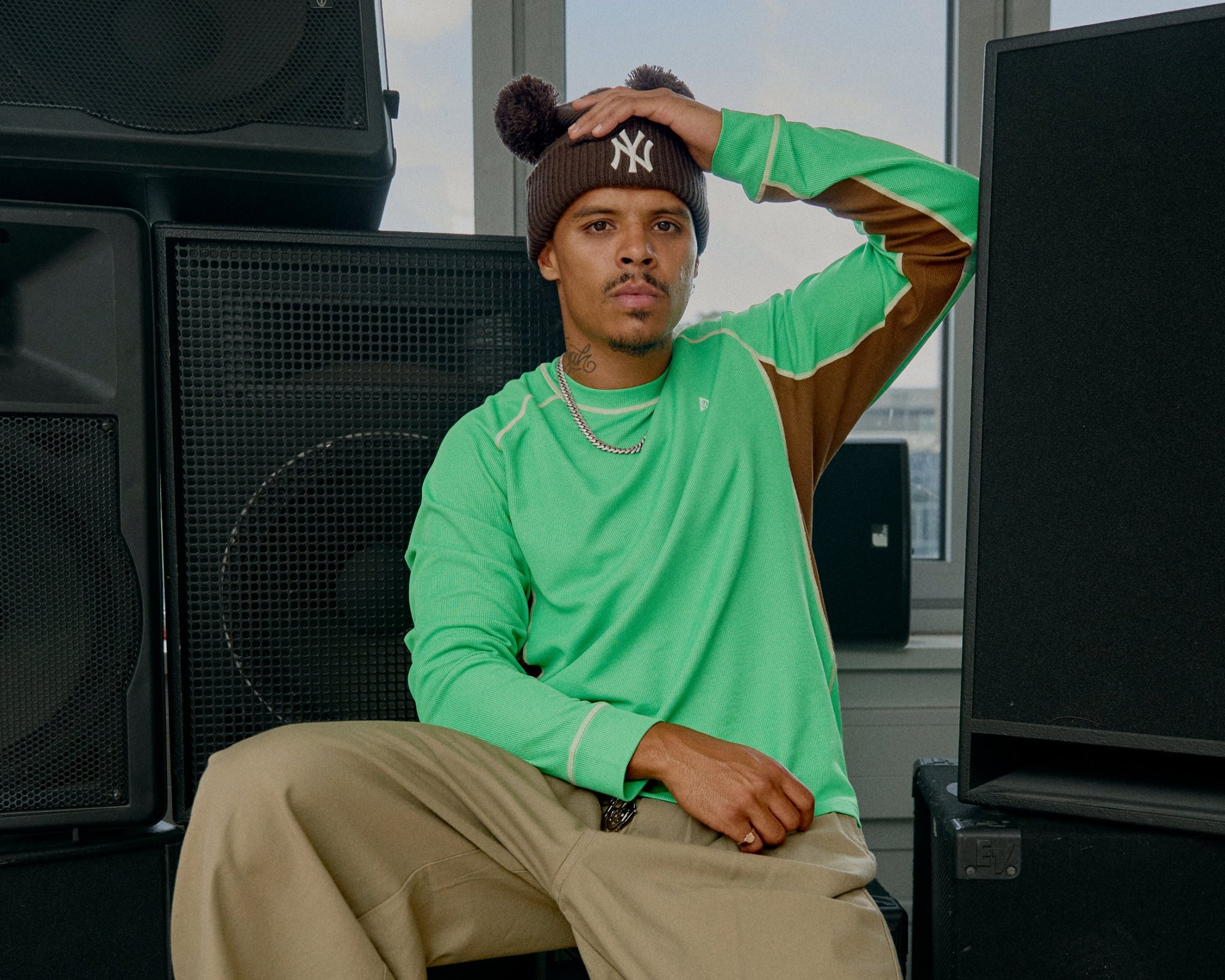 Chemise verte à manches longues avec des accents marron et des coutures claires, associée à un pantalon beige clair. Un bonnet marron avec des pompons repose sur la tête.