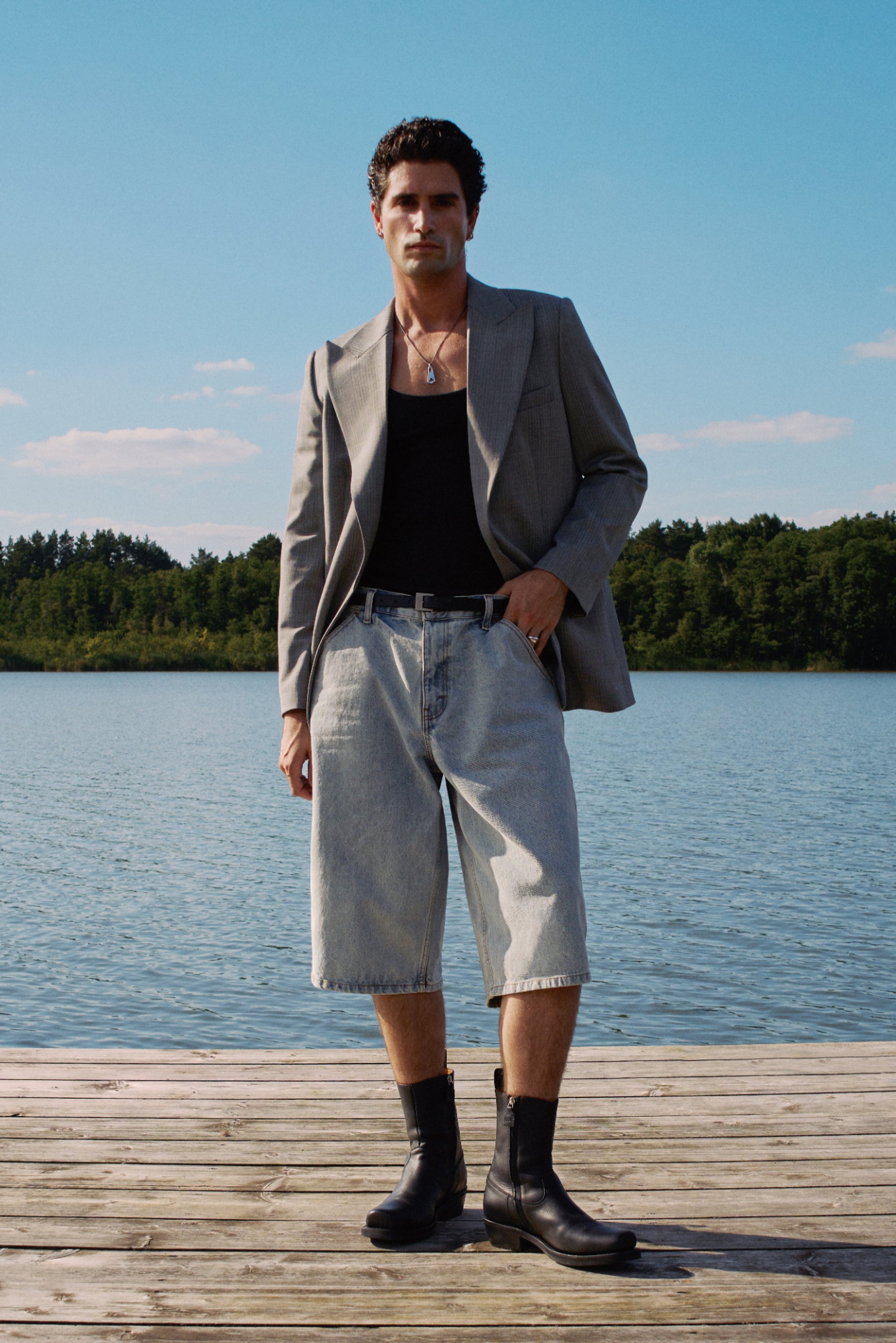 Man stands on a wooden dock by a lake, wearing a grey blazer, black tank top, loose denim shorts, and black ankle boots, against a blue sky.