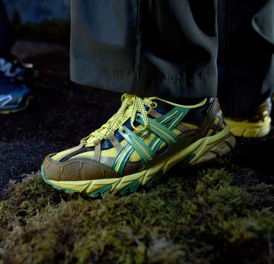Close-up of two pairs of athletic shoes on a mossy surface. One pair is bright yellow and brown; the other is black and gray.
