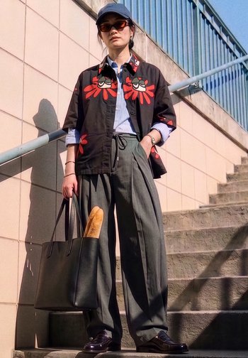 Persona con camisa negra con diseños de flores rojas, pantalones anchos grises, gafas de sol y gorra, sosteniendo un bolso tote negro con una baguette cerca de unas escaleras.
