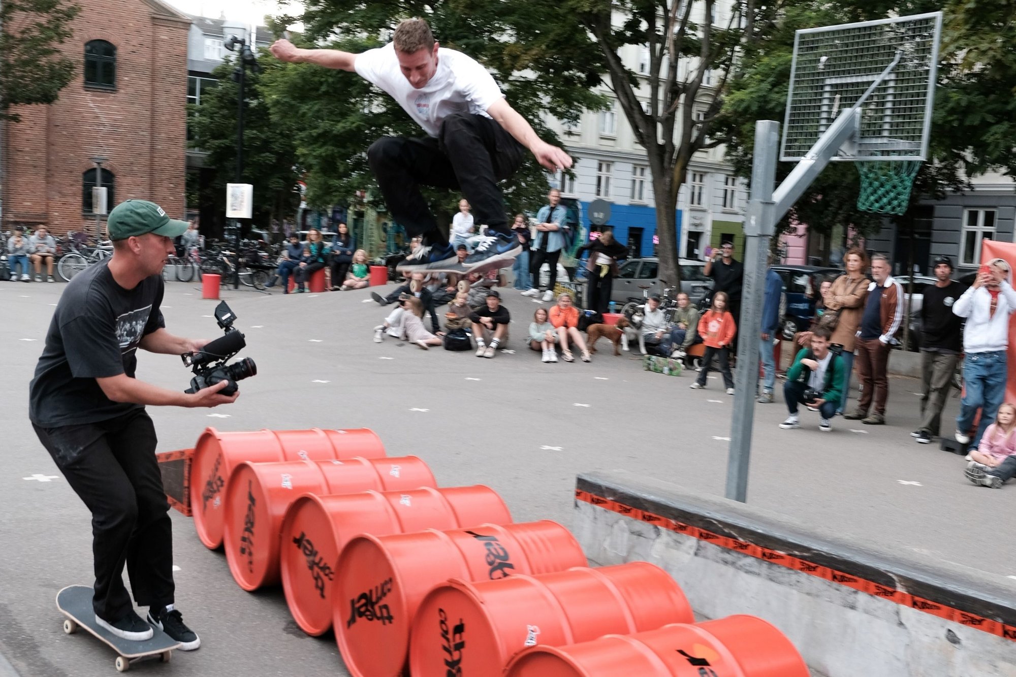 Skateboardåkare i luften ovanför orange fat, iklädd en vit t-shirt och svarta byxor, med en publik som tittar i en parkmiljö.