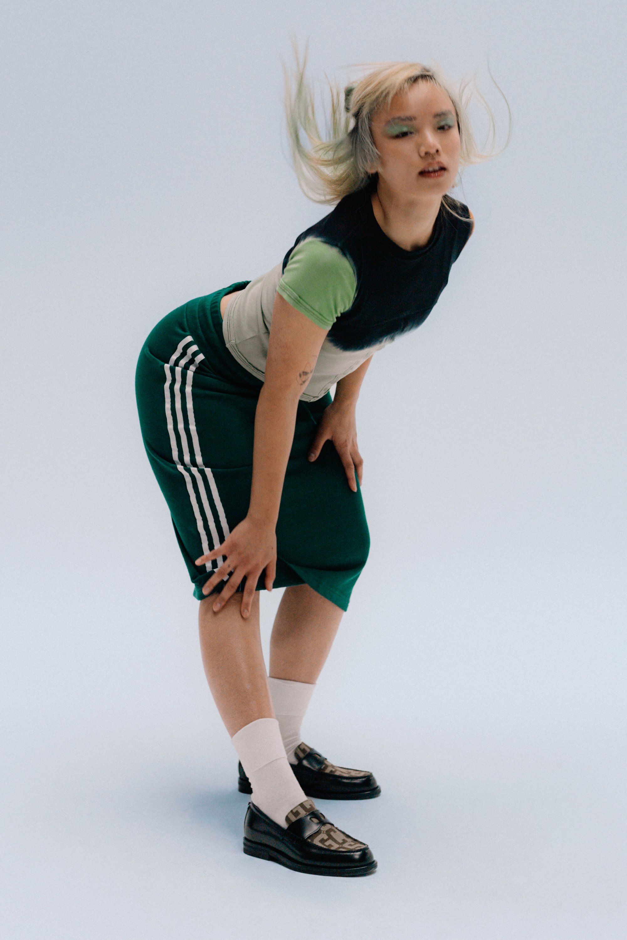 Una persona con cabello rubio claro, vestida con un conjunto negro y verde y zapatos con estampado, se inclina ligeramente hacia adelante contra un fondo pálido.