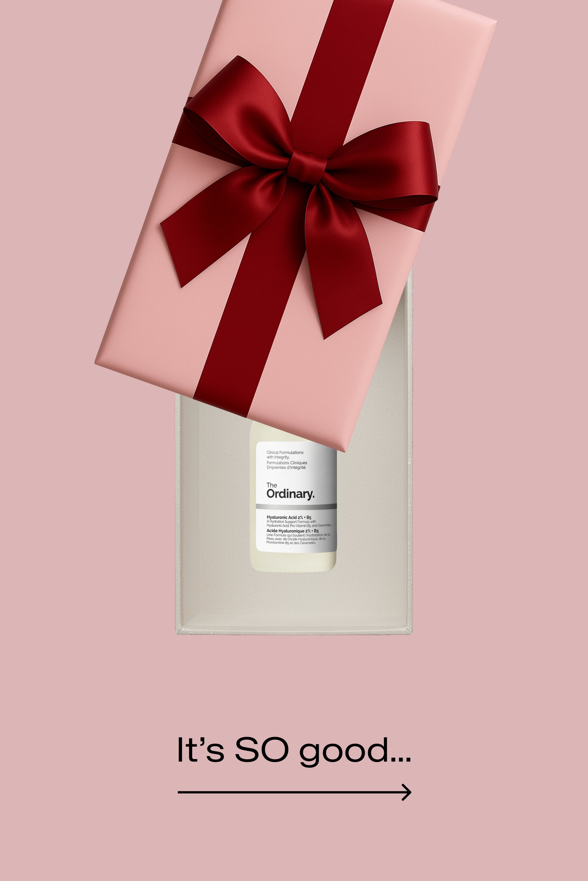 Caja de regalo rosa con un lazo de raso rojo. Dentro, una botella transparente etiquetada "The Ordinary" con texto en blanco y negro, que contiene suero de ácido hialurónico.
