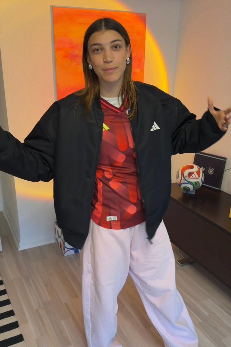 Young person in a red sports jersey and black jacket stands with arms open inside a room with a soccer ball and orange artwork in the background.