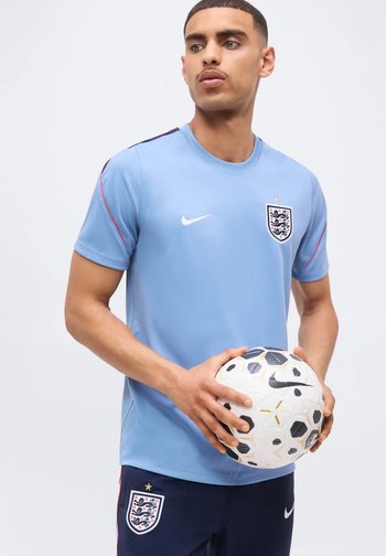 Homme portant un maillot de football bleu clair de l'Angleterre et un short bleu marine tenant un ballon de football Nike blanc et noir sur un fond uni.