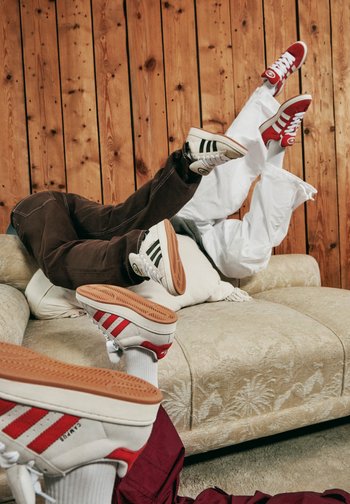 Füße in stylischen Sneakern mit kontrastierenden Farben, verspielt auf einer Couch vor einem Holz-Hintergrund positioniert. Gemütliche Atmosphäre.