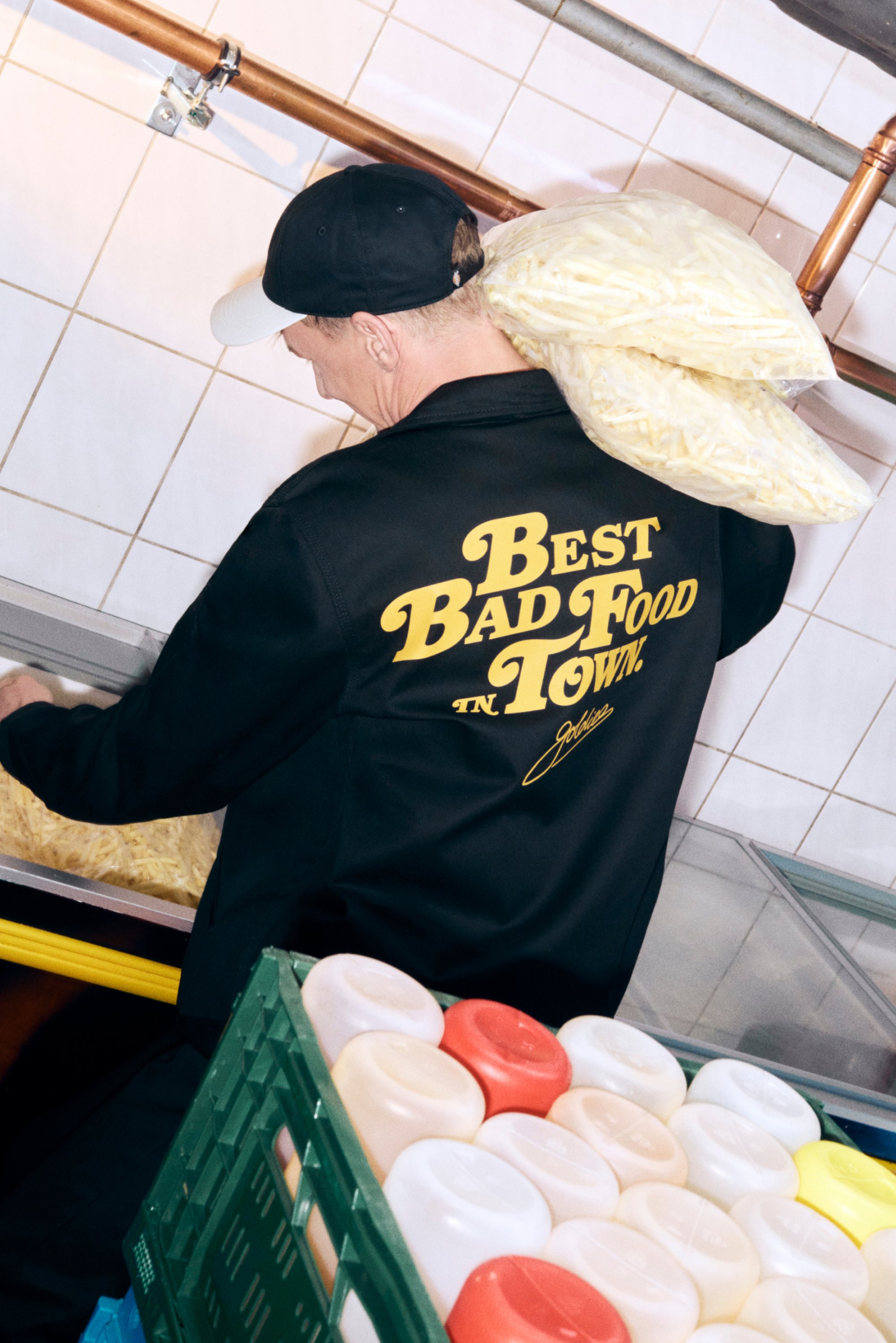 Eine Person in einer schwarzen Jacke mit gelbem Text trägt Tüten mit Pommes frites in einer Küche, während im Vordergrund bunte Behälter sichtbar sind.
