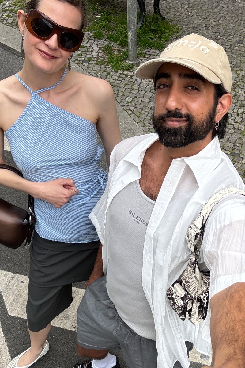 Due persone sorridono in una strada della città. La donna indossa una maglietta a righe blu e l'uomo sfoggia una camicia chiara e un cappellino, mentre tiene in mano una borsa piccola.
