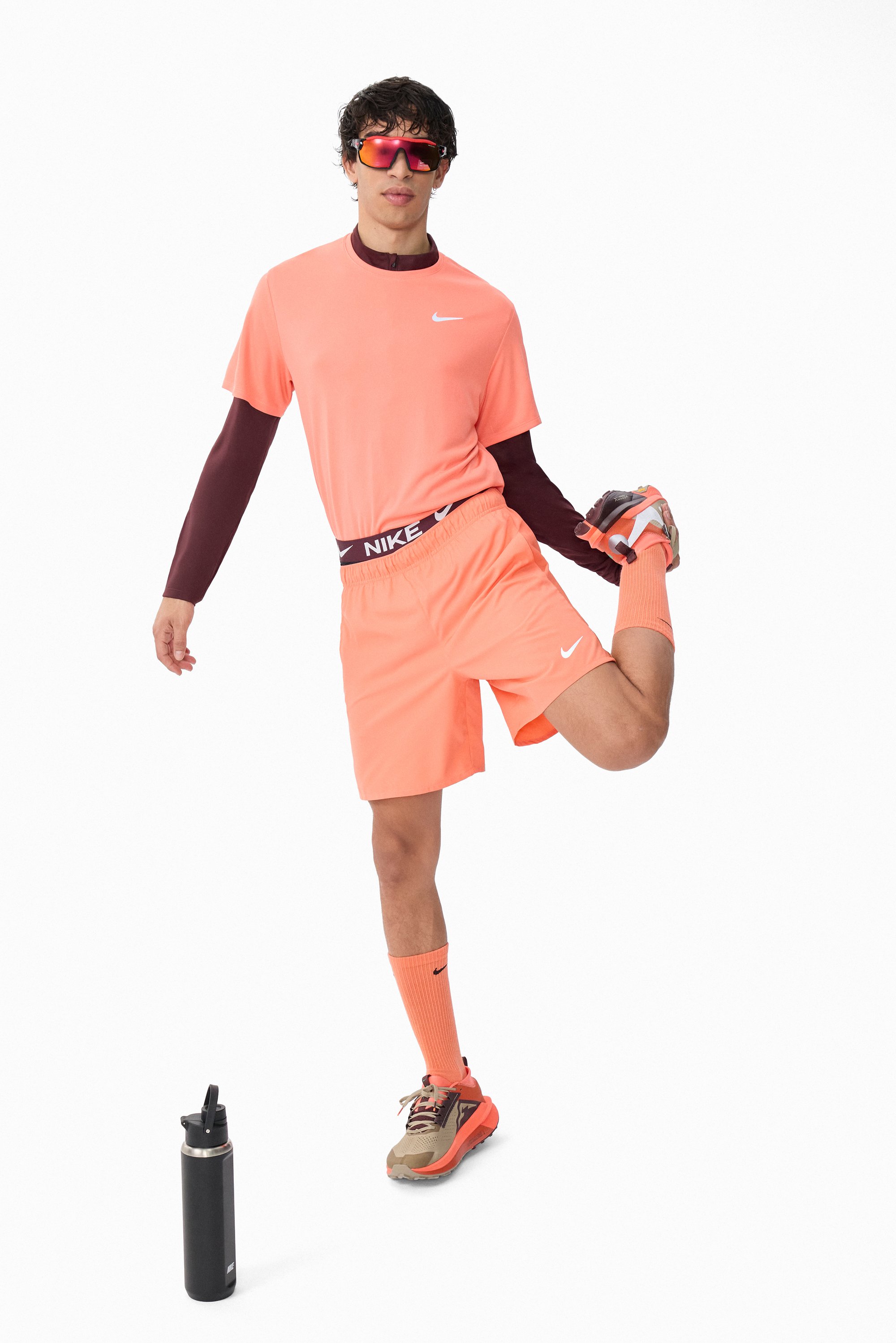 Camiseta y pantalones cortos deportivos de color naranja neón con una cinturilla negra de Nike. Calcetas naranja hasta la rodilla, zapatillas gray y naranja, y una botella de agua negra.
