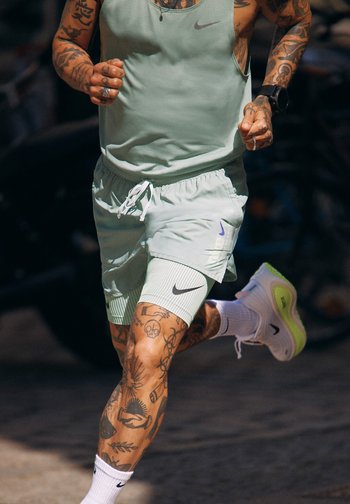 Tatoveret person iført lysegrønt sportstøj og hvide sneakers løber udendørs; kun kroppen fra skuldre til lægge er synlig.