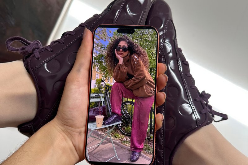 Hand, die ein Telefon hält, auf dem eine Frau mit lockigem Haar, Sonnenbrille, brauner Jacke und rosa Hose zu sehen ist, die draußen neben einem Fahrrad und einem Tisch mit einem eisgekühlten Getränk posiert, dabei dunkellila Schuhe trägt.