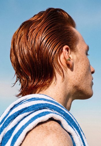 Giovane con capelli rossi bagnati pettinati all'indietro, che indossa un asciugamano a righe sulla spalla, guarda a destra contro un cielo azzurro limpido.
