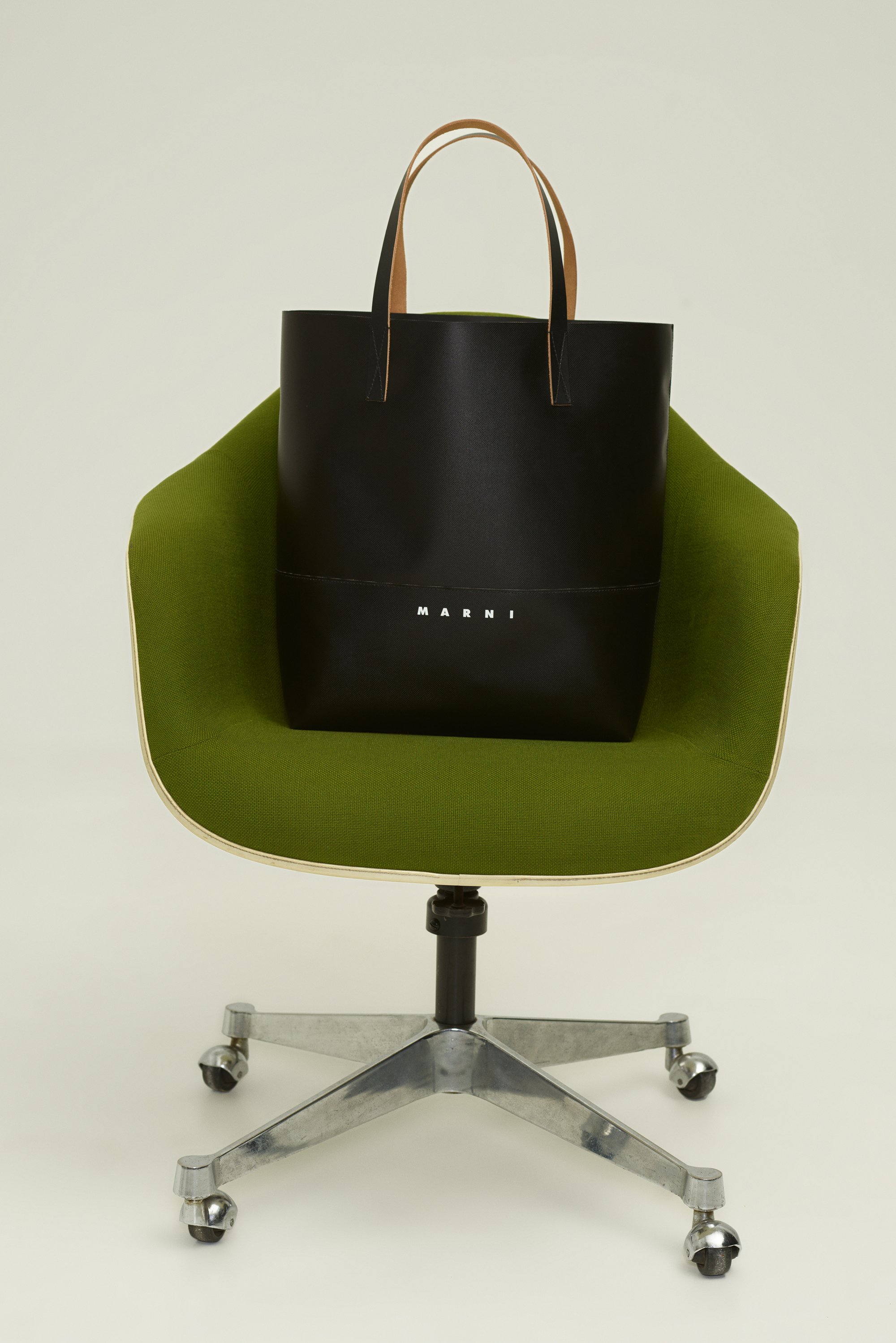 Bolso tote de cuero negro con asas marrones, que presenta una costura horizontal y el logo blanco "MARNI" en la parte inferior, colocado sobre una silla verde.