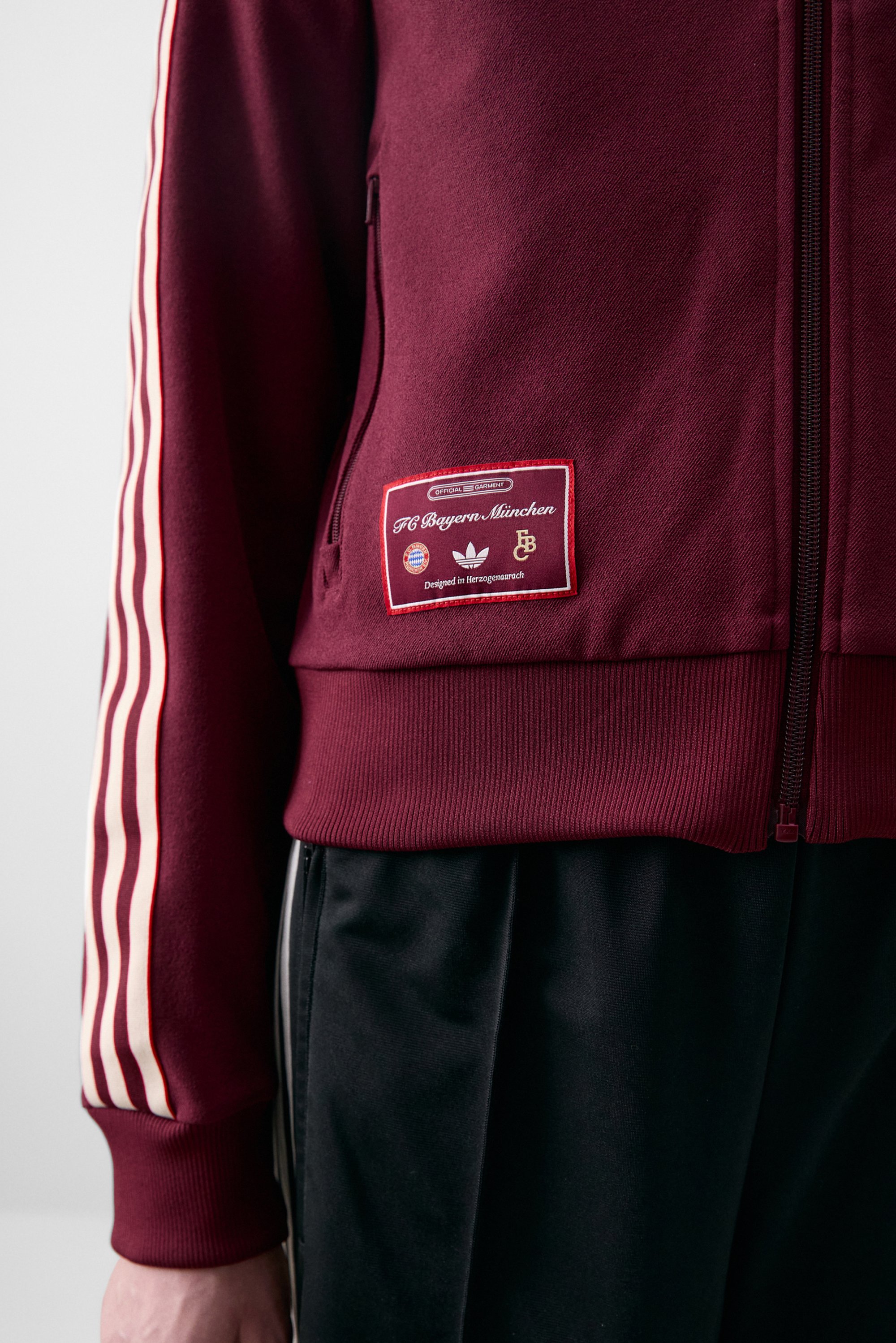 Bordeauxrode zip-up jack met FC Bayern München branding en drie witte strepen op de mouwen, gecombineerd met zwarte broek.