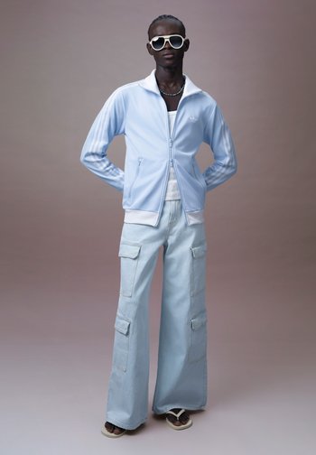 Persoon die een lichtblauw Adidas trainingsjack, wijde cargobroek, wit hemdje, grote zonnebril en sandalen draagt tegen een neutrale achtergrond.