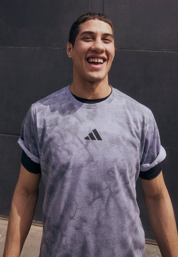 Un jeune homme souriant dans un t-shirt de sport léger violet se tient contre un mur sombre, dégageant confiance et énergie.