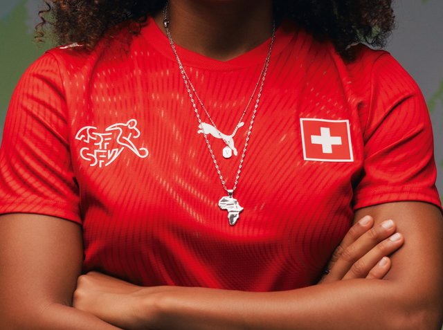 Person in einem roten Sporttrikot mit Schweizer Flagge und Puma-Logos, trägt zwei silberne Halsketten, Arme vor der Brust verschränkt.