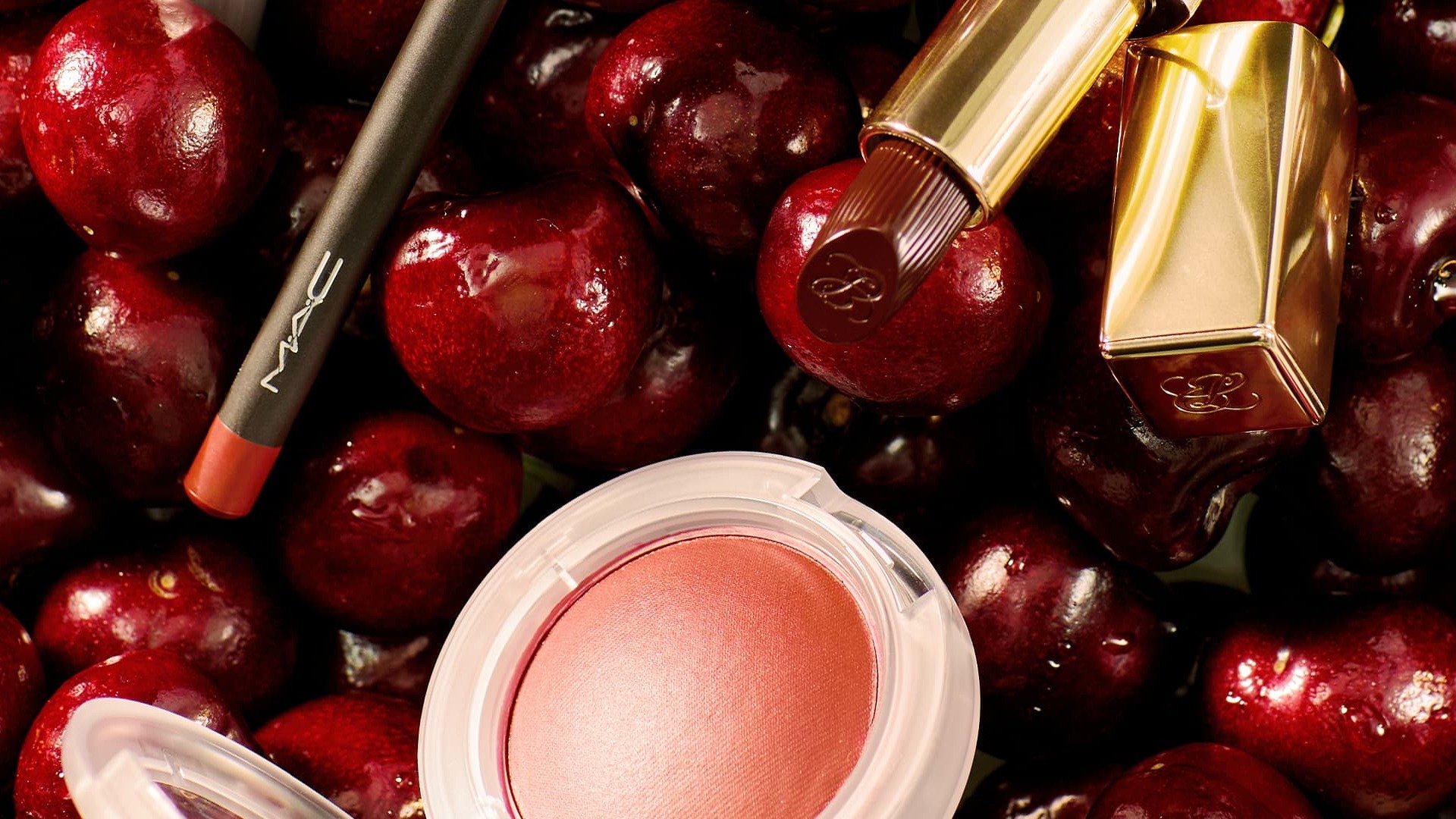 Crayon à lèvres rouge cerise, rouge à lèvres et compact de blush pêche posés sur un lit de cerises fraîches rouge foncé.