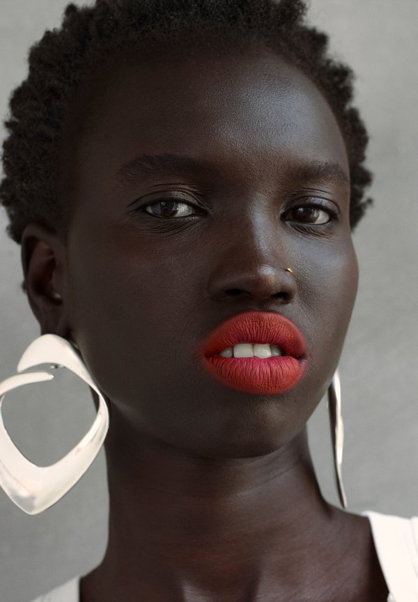 Gros plan sur une femme à la peau foncée, avec des cheveux courts et bouclés, du rouge à lèvres rouge vif, un anneau au nez et de grandes boucles d'oreilles géométriques en argent, sur un fond neutre.
