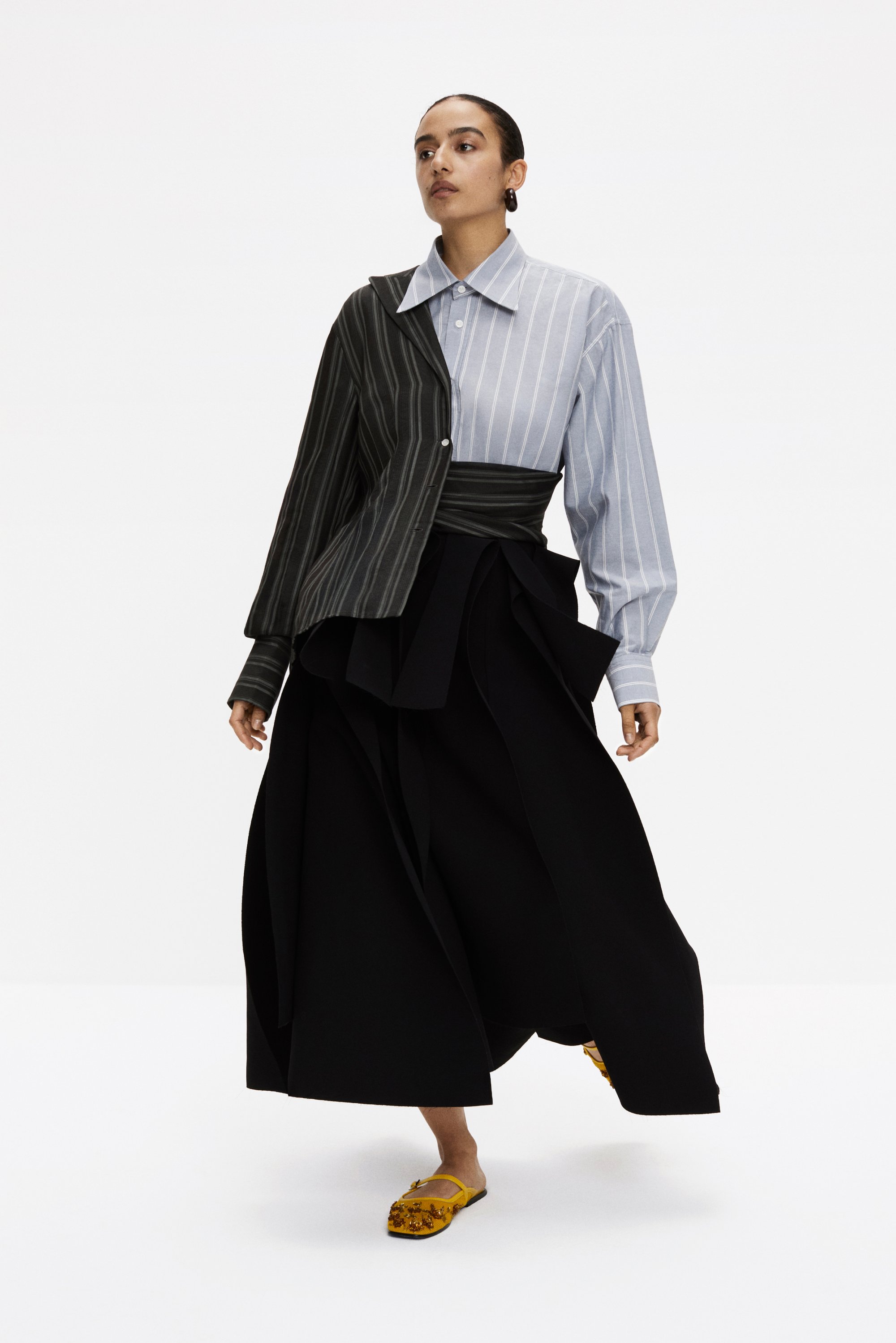 Una modella cammina con sicurezza, indossando un outfit unico e asimmetrico composto da una camicia a righe e una gonna nera fluente, abbinato a scarpe gialle.
