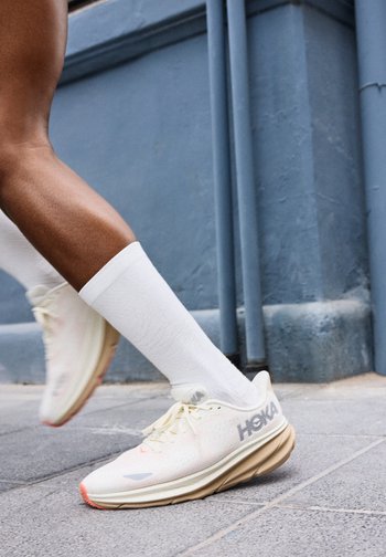 Άτομο με λευκές κάλτσες και λευκά παπούτσια τρεξίματος Hoka, που απεικονίζεται σε μέση βήματος σε πεζοδρόμιο με γκρι πλακάκια δίπλα σε μπλε τοίχο.