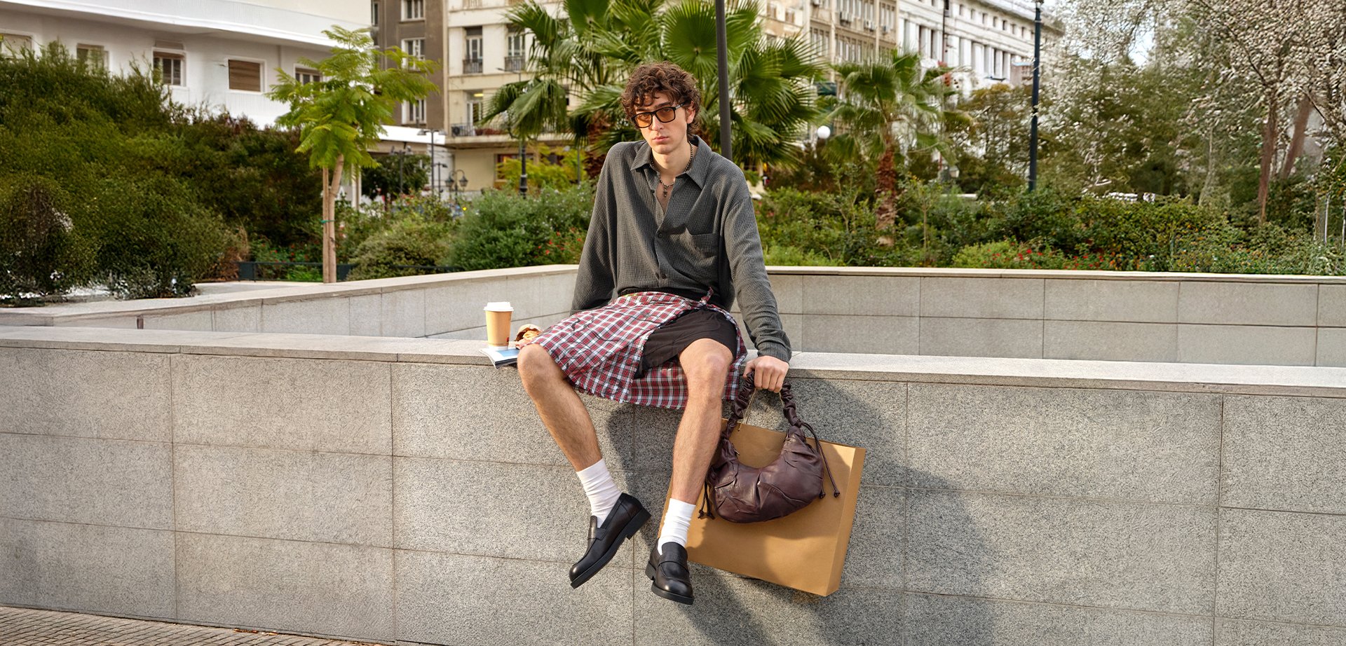 Joven con pelo rizado sentado en un saliente de piedra en un parque urbano, sosteniendo una bolsa marrón y un vaso de café para llevar cerca.